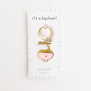 May include: Gold-colored keychain featuring a pink bowl of noodles with a smiling face. The keychain has a gold-colored ring and a small gold-colored chopstick. The text "it's a keychain!" is printed on a white card behind the keychain. The text "a jar of pickles" is printed on the bottom of the card.