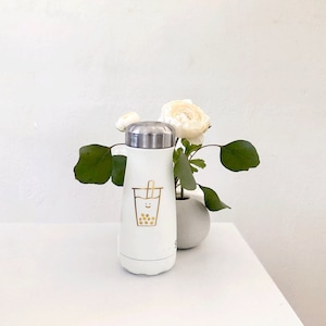 Op de afbeelding: Witte geïsoleerde waterfles met een zilveren deksel en een gouden bubble tea-afbeelding. De fles staat op een wit oppervlak met een kleine vaas met witte bloemen en groene bladeren.
