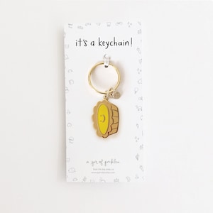 May include: Gold keychain shaped like a pie with a smiling face. The keychain is attached to a white card with the text "it's a keychain!" and "a jar of pickles" printed on it.