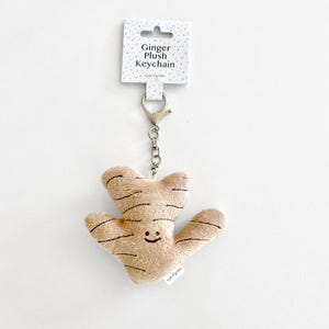 Pode incluir: Um chaveiro de gengibre de pelúcia marrom com um rosto sorridente. O chaveiro é preso a uma corrente de metal prateada e tem uma etiqueta que diz "Ginger Plush Keychain" e "a jar of giggles".