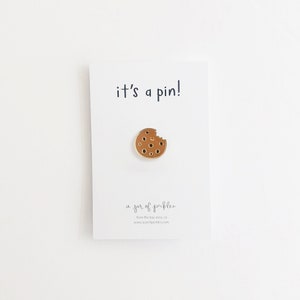 May include: A brown enamel pin in the shape of a bitten cookie with a smiling face. The pin is on a white card that reads "it's a pin!" and "a jar of pickles."