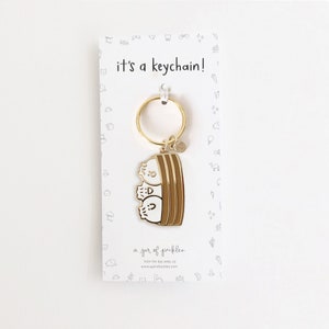May include: Gold-colored keychain featuring three cartoon ghost characters in a brown bowl. The keychain is attached to a gold-colored ring. The card reads "it's a keychain!" and "a jar of pickles".