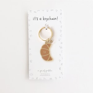 May include: A gold-coloured keychain with a brown croissant charm. The charm has a smiley face and the text "it's a keychain!" is printed on the card.