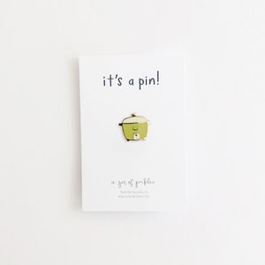 May include: A green and gold enamel pin of a rice cooker with a smiling face. The pin says "it's a pin!" and "a jar of pickles" with a website address.