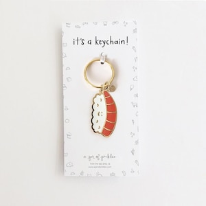 May include: Gold keychain with a red and white sushi charm. The charm has a smiling face and the text "it's a keychain!" is printed on the card.