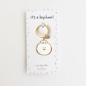 May include: Gold keychain featuring a white cartoon dumpling with a happy face. The keychain is packaged on a white card with the text "it's a keychain!" and "a jar of pickles" printed on it.
