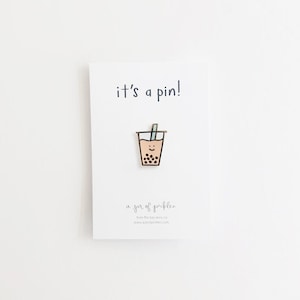 Pode incluir: Um alfinete de esmalte dourado com uma ilustração de desenho animado de uma xícara de chá de bolhas com um canudo e um rosto sorridente. O texto "it's a pin!" é impresso acima da imagem e "a jar of pickles." é impresso abaixo da imagem.