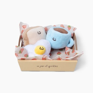 May include: A breakfast-themed gift set with plush toys of toast, a fried egg, and a coffee cup, each with a smiling face. The items are in a small, light brown box lined with strawberry-printed paper. The box has the text "a jar of pickles".