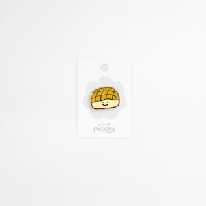 Puede incluir: Un pin de esmalte metálico con forma de pan de melón amarillo y crema con una cara sonriente sencilla. El pin está sujeto a una tarjeta de presentación rectangular blanca con el texto a jar of pickles impreso en gris claro en la parte inferior.