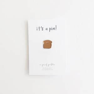 May include: A pin featuring a smiling piece of toast. The pin is brown with a gold outline and is attached to a white card that reads "it's a pin!" and "a jar of pickles."
