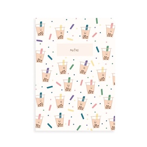 Könnte beinhalten: Ein Notizbuch mit weißem Einband, das ein Muster aus lächelnden Bubble-Tea-Bechern in transparenten Plastikbechern mit bunten Strohhalmen zeigt. Das Wort "Notizen" ist auf einem hellrosa Rechteck in der Mitte aufgedruckt. Bunte Konfetti punkten den Hintergrund.
