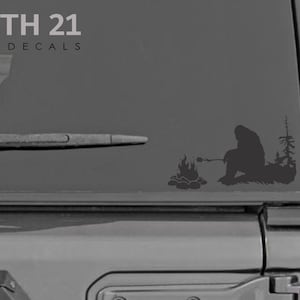 May include: A black and white vinyl decal of a Bigfoot sitting by a campfire, roasting a marshmallow. The decal is on the back window of a gray vehicle. The text "NORTH 21 VINYL DECALS" is at the top of the image.