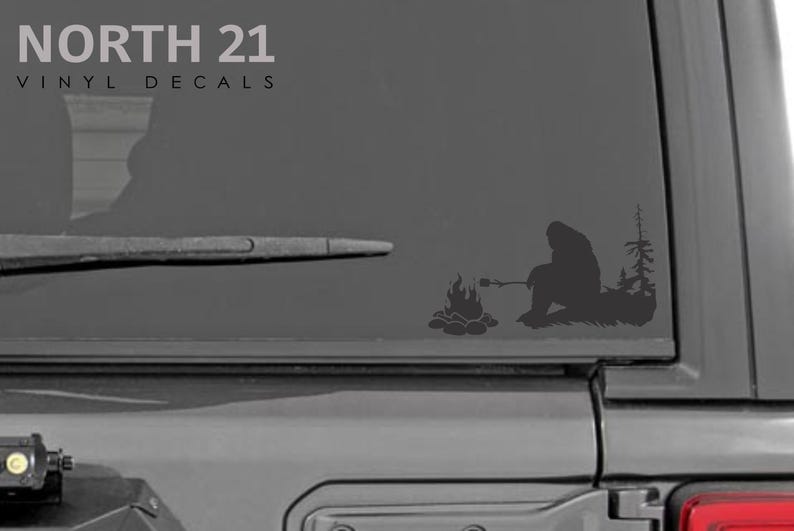 May include: A black and white vinyl decal of a Bigfoot sitting by a campfire, roasting a marshmallow. The decal is on the back window of a gray vehicle. The text "NORTH 21 VINYL DECALS" is at the top of the image.