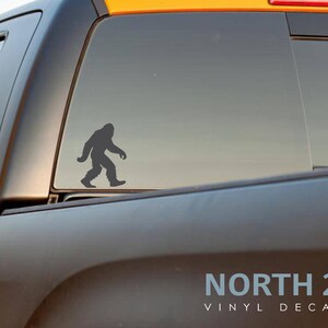 May include: A gray vinyl decal of a Bigfoot silhouette walking to the right is affixed to the rear window of a black truck. The decal is positioned on the window's lower half. The text "NORTH 21 VINYL DECALS" is visible at the bottom.