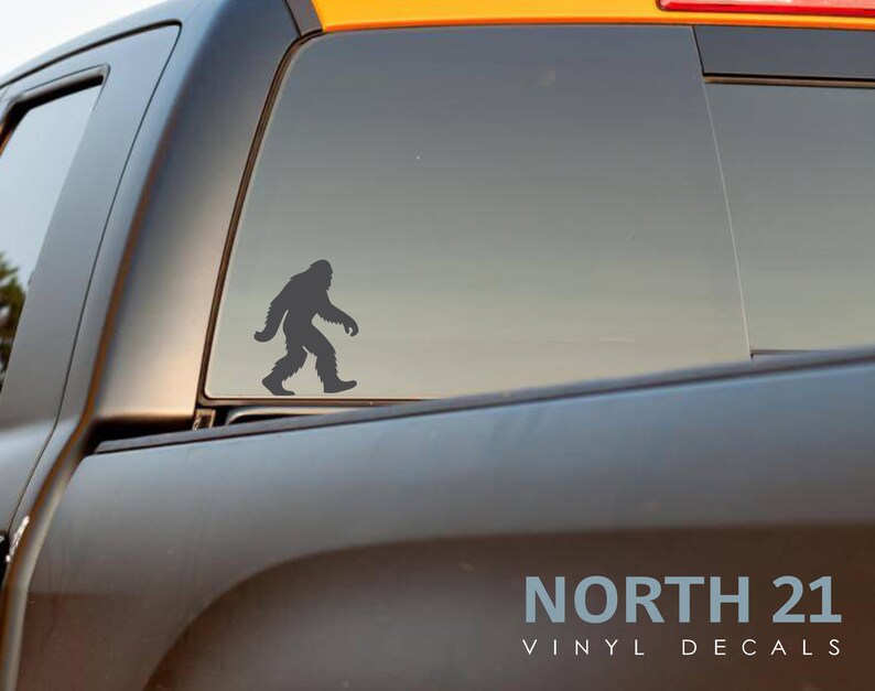 May include: A gray vinyl decal of a Bigfoot silhouette walking to the right is affixed to the rear window of a black truck. The decal is positioned on the window's lower half. The text "NORTH 21 VINYL DECALS" is visible at the bottom.
