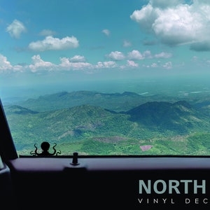 May include: A black octopus vinyl decal with the text "NORTH 21 VINYL DECALS" on a black background. The decal is placed on the back window of a car, with a view of a mountain range and a blue sky with white clouds.