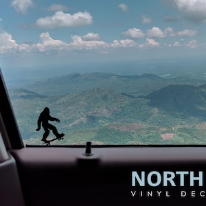 May include: A black silhouette of a Bigfoot character skateboarding, with the text "NORTH 21 VINYL DECALS" below. The image is taken from inside a car looking out the window at a mountain range.