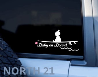 May include: White vinyl decal of a baby girl sitting on a surfboard with the text "Baby on Board" and a pink heart.