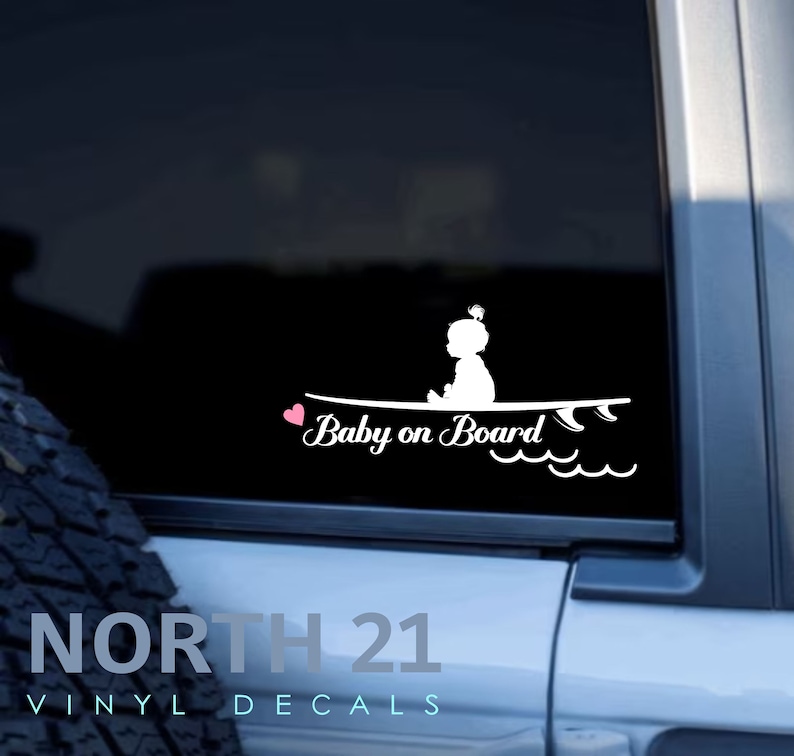 May include: White vinyl decal of a baby girl sitting on a surfboard with the text "Baby on Board" and a pink heart.