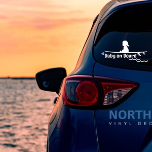 May include: A blue car with a white vinyl decal on the back window that says "Baby on Board" with a silhouette of a child sitting on a surfboard. The car is parked in front of a body of water with a sunset in the background.