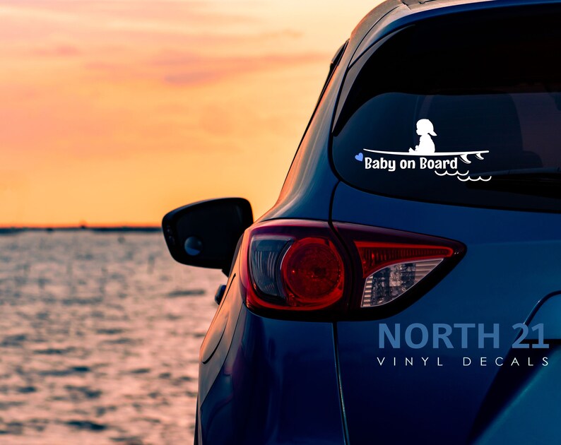 May include: A blue car with a white vinyl decal on the back window that says "Baby on Board" with a silhouette of a child sitting on a surfboard. The car is parked in front of a body of water with a sunset in the background.