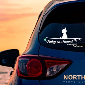 May include: White vinyl decal with a silhouette of a baby sitting on a surfboard with the text "Baby on Board" and a small green heart. The decal is on the back window of a blue car.