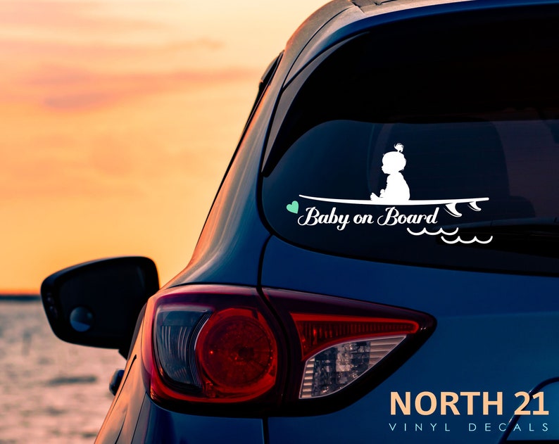 May include: White vinyl decal with a silhouette of a baby sitting on a surfboard with the text "Baby on Board" and a small green heart. The decal is on the back window of a blue car.