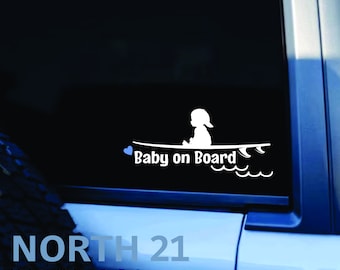 May include: White vinyl decal with a silhouette of a baby sitting on a surfboard with the text "Baby on Board" and a blue heart. The decal is on a black car window.