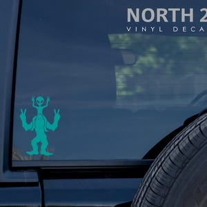 May include: Teal vinyl decal of a Bigfoot holding a small alien on its shoulders, both with peace signs. The decal is on the back window of a black car. The text "NORTH 21 VINYL DECALS" is at the top of the image.