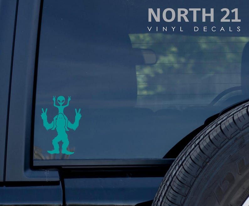 May include: Teal vinyl decal of a Bigfoot holding a small alien on its shoulders, both with peace signs. The decal is on the back window of a black car. The text "NORTH 21 VINYL DECALS" is at the top of the image.