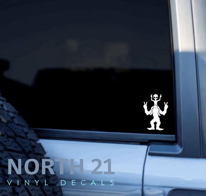 May include: White vinyl decal of a Bigfoot with an alien riding on his shoulders. The decal is on the back window of a car. The text "NORTH 21 VINYL DECALS" is visible on the car.