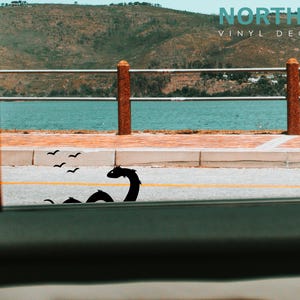 May include: A black vinyl decal of a sea serpent with a long neck and a small head, with a flock of birds flying above it. The decal is placed on a gray asphalt road with a yellow line down the center. The road is next to a body of water with a green hill in the background. The text "NORTH 21 VINYL DECALS" is visible in the top right corner of the image.