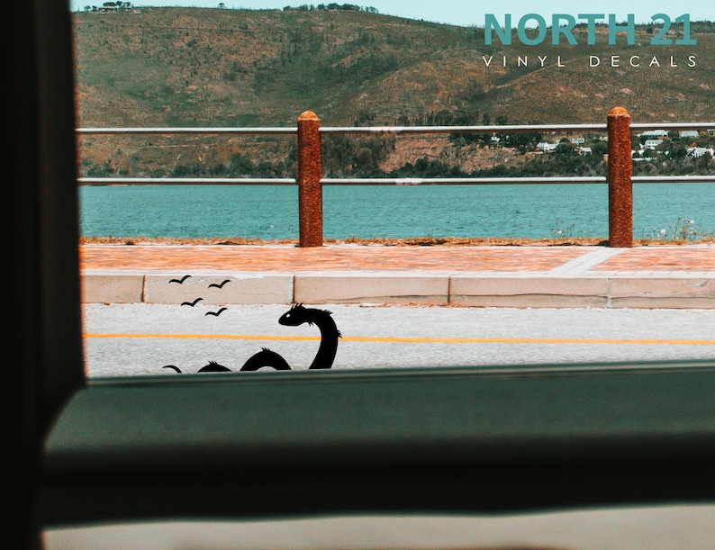 May include: A black vinyl decal of a sea serpent with a long neck and a small head, with a flock of birds flying above it. The decal is placed on a gray asphalt road with a yellow line down the center. The road is next to a body of water with a green hill in the background. The text "NORTH 21 VINYL DECALS" is visible in the top right corner of the image.