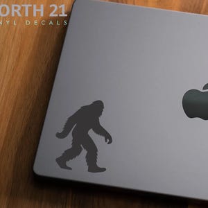 May include: A gray laptop with a black Bigfoot decal and a black Apple logo. The Bigfoot silhouette is walking to the left. The text "NORTH 21 VINYL DECALS" is in the upper left corner.