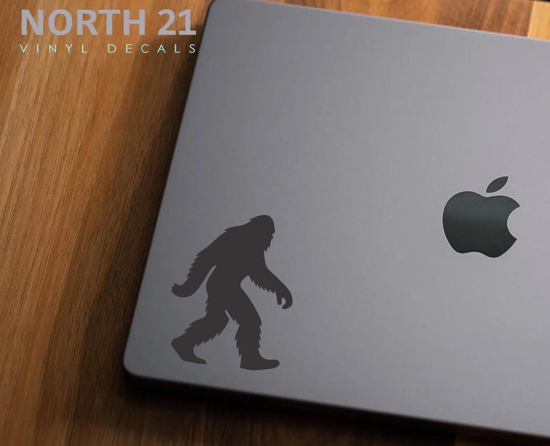 May include: A gray laptop with a black Bigfoot decal and a black Apple logo. The Bigfoot silhouette is walking to the left. The text "NORTH 21 VINYL DECALS" is in the upper left corner.