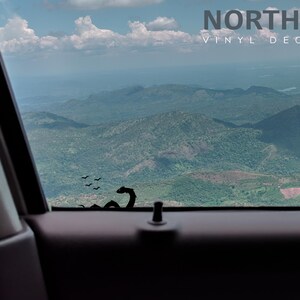 May include: A black vinyl decal of a dragon with birds flying above it, placed on a car window with a view of a mountain range in the distance.