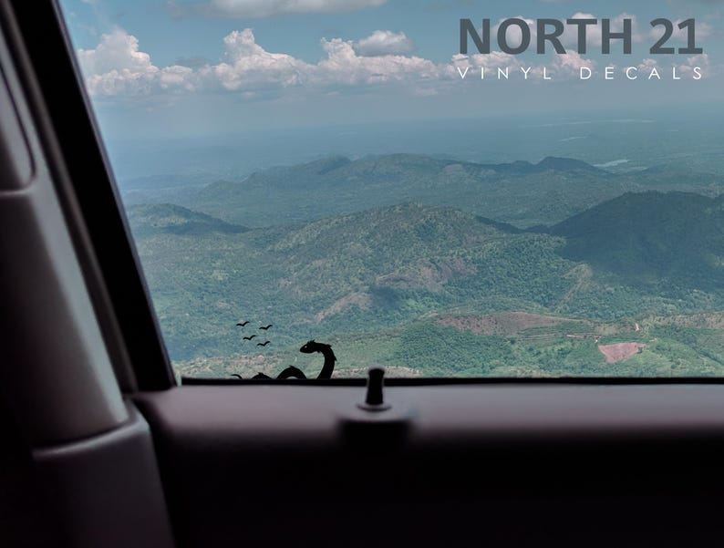 May include: A black vinyl decal of a dragon with birds flying above it, placed on a car window with a view of a mountain range in the distance.
