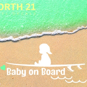 May include: White silhouette of a baby sitting on a surfboard with the text "Baby on Board" and a green heart on the left side of the surfboard. The background is a sandy beach with a turquoise ocean.