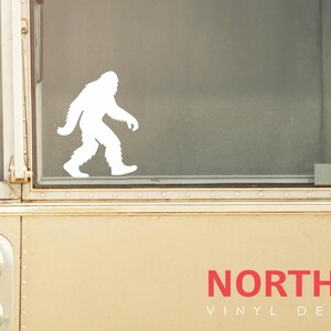 May include: A white vinyl decal of a Bigfoot silhouette walking to the left is displayed on a window. The decal is set against a neutral background. The text "NORTH 21 VINYL DECALS" is printed in red.