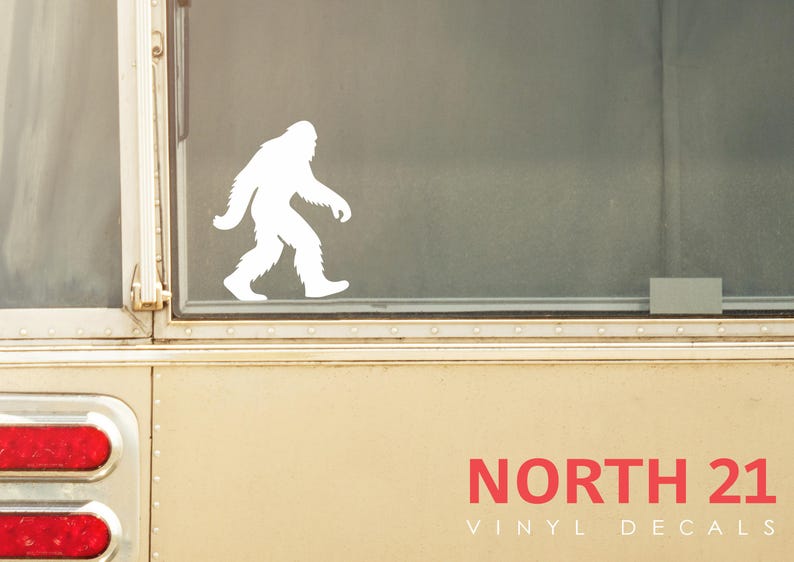May include: A white vinyl decal of a Bigfoot silhouette walking to the left is displayed on a window. The decal is set against a neutral background. The text "NORTH 21 VINYL DECALS" is printed in red.