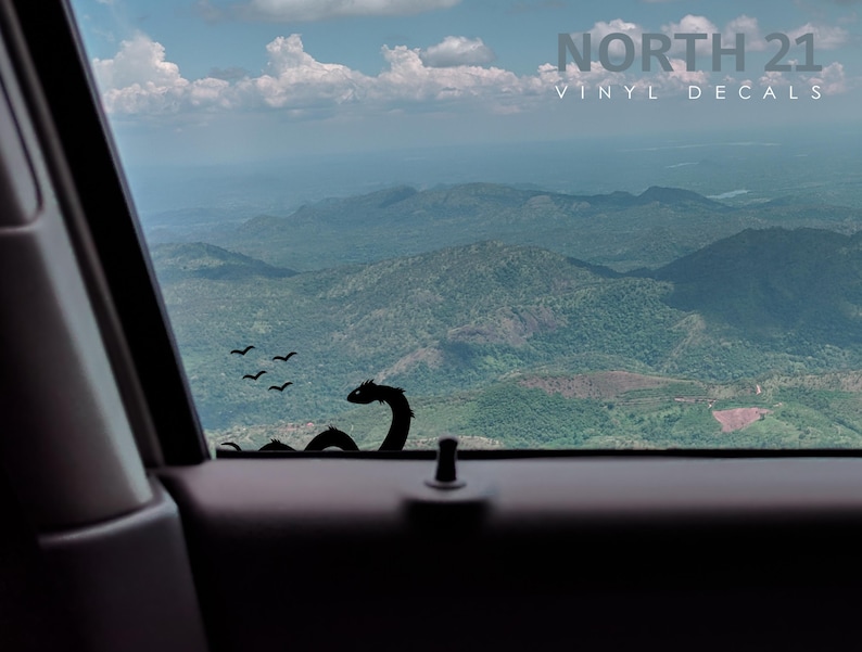 May include: A black vinyl decal of a sea serpent with a long neck and a small head. The decal is placed on the back window of a car. The car is driving through a mountainous landscape. The decal is part of a set of vinyl decals sold by North 21.