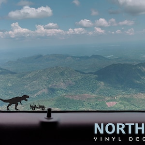 May include: A black silhouette of a Tyrannosaurus Rex and a jeep driving through a mountainous landscape. The image is a vinyl decal sticker with the text "NORTH 21 VINYL DECALS" at the bottom.