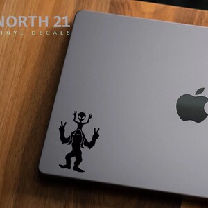 May include: Black and white vinyl decal of a Bigfoot carrying an alien on its shoulders, both figures are making the peace sign. The decal is on a gray laptop with the Apple logo.