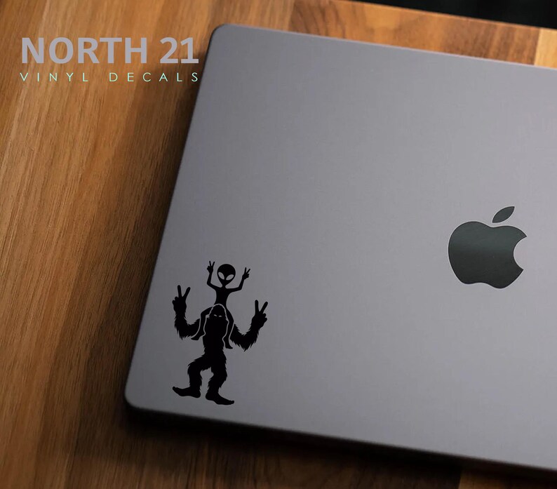 May include: Black and white vinyl decal of a Bigfoot carrying an alien on its shoulders, both figures are making the peace sign. The decal is on a gray laptop with the Apple logo.