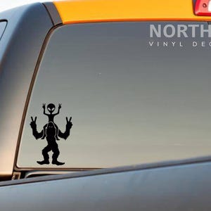 May include: Black vinyl decal of a Bigfoot with an alien riding on his shoulders, both giving the peace sign. The decal is on the back window of a black truck. The text "NORTH 21 VINYL DECALS" is in white text at the top of the window.