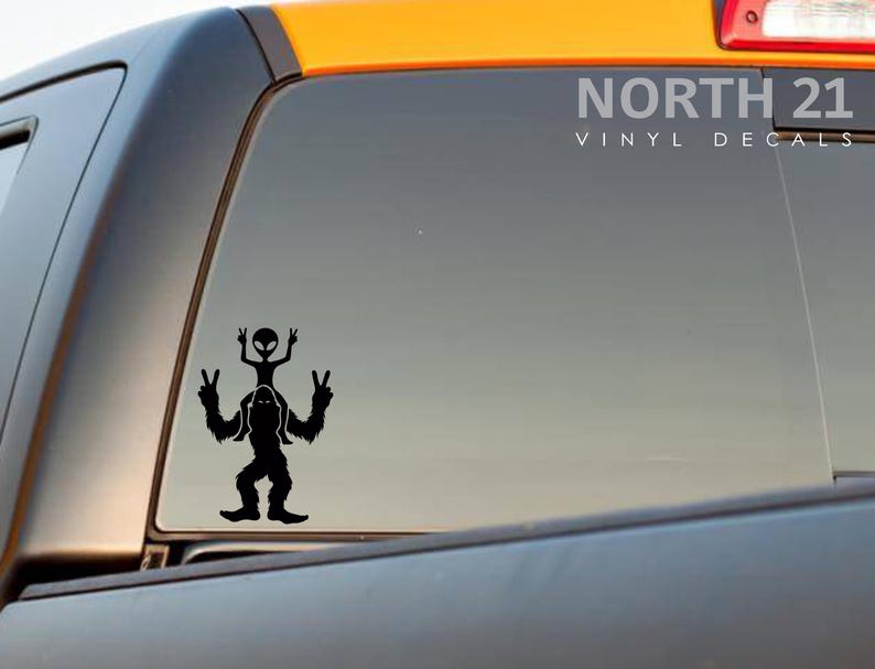 May include: Black vinyl decal of a Bigfoot with an alien riding on his shoulders, both giving the peace sign. The decal is on the back window of a black truck. The text "NORTH 21 VINYL DECALS" is in white text at the top of the window.