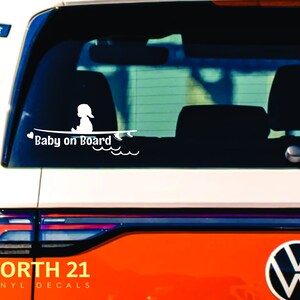 May include: White vinyl decal with a silhouette of a baby sitting on a surfboard with the text "Baby on Board" and a heart. The decal is on the back window of a red car.