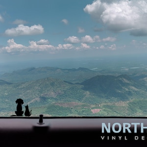 May include: A black silhouette of a dog and cat sitting side by side, looking out of a car window. The view from the window shows a mountainous landscape with green hills and a blue sky with white clouds. The text "NORTH 21 VINYL DECALS" is visible at the bottom of the image.