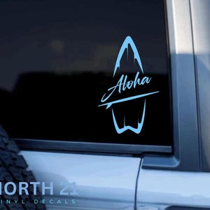 May include: A blue surfboard decal with the word "Aloha" written on it. The decal is on a black background and is attached to a car window.