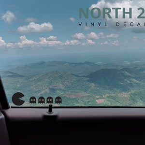 Puede incluir: Una ventana de coche con vista a montañas y nubes. Un Pac-Man negro y cuatro fantasmas son visibles en la ventana. El texto "NORTH 21 VINYL DECALS" está en la parte superior de la imagen.